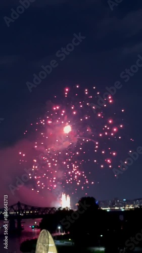 Vibrant red fireworks burst over city river at night, creating magical sky spectacle. dazzling celebration lights up skyline near bridge, drawing attention to urban festivity.
