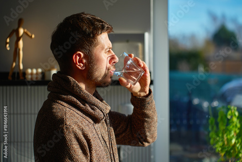 Photography Adult man drinking fresh clean water from glass while standing in kitchen near window with sunlight