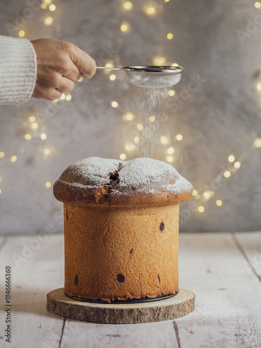A woman's hand sprinkles powdered sugar onto a panettone. Christmas lights with a bokeh effect in the background.