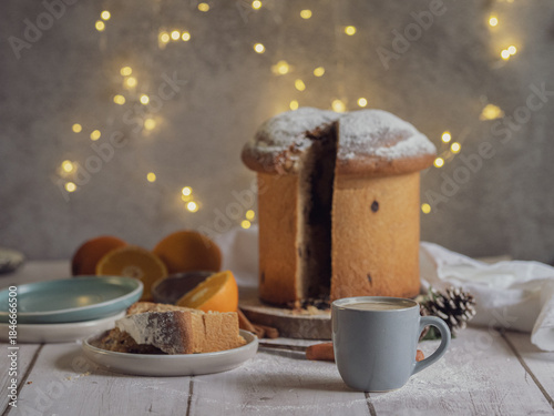 A slice of traditional Italian panettone next to a cup of coffee on a wooden table with Christmas lights. Space for text
