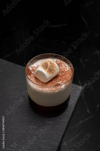 A close-up shot of an elegant cocktail garnished with a marshmallow, placed on a dark surface, evokes luxury. The drink features a marshmallow topping and brown sprinkles.