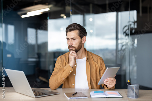 Bilde på lerret Man in smart casual attire at a modern office desk, holding a tablet and reviewi