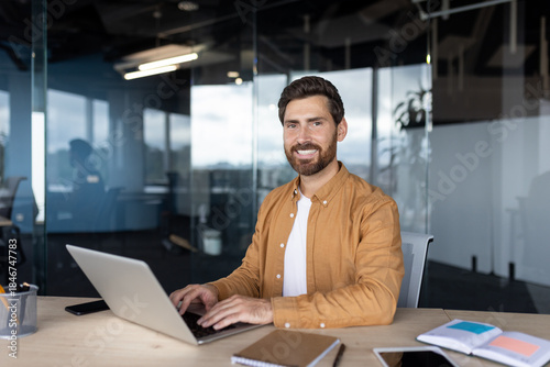 Businessman with a beard, wearing an orange shirt, working on a laptop at his desk, smiling at the camera in a contemporary office setting with glass partitions