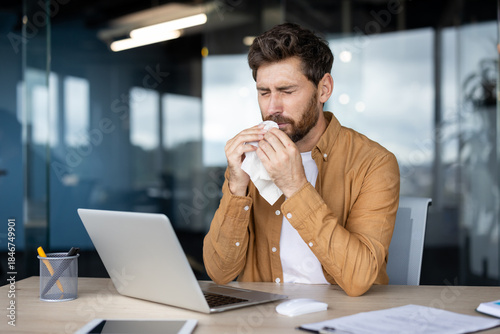 Фотография Man blowing his nose into a tissue at his office desk while working on a laptop,