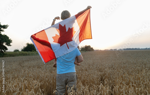 large Canadian flag draped over boy sitting on shoulders of man, standing in a wheat field at sunset. Happy Canada Day Pride, freedom, patriotism. A journey across the country. A national symbol