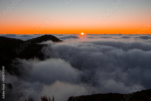 Canvas-taulu The sun peeks above the horizon, casting an orange band as mountain ridgelines pierce clouds near Pico do Arieiro and Pico Ruivo in Madeira under soft diffuse light