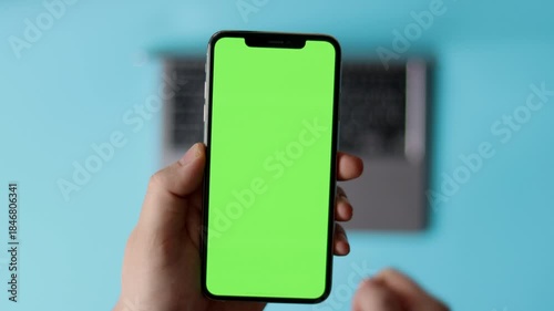 Person holding smartphone with green screen in front of laptop. Flatlay. Mock up concept, green, blank screen