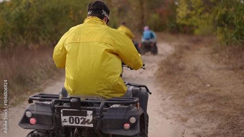 rear view rider yellow jacket atv trail kicking up dust along rural dirt path through autumn forest, helmeted adult navigating winding track with friends ahead,