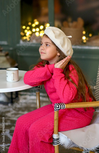 Adorable little girl in a festive winter setting, sitting on a vintage bench and holding a warm cup. Child wearing a bright pink cozy outfit and a white beret, smiling gently. New Year concept