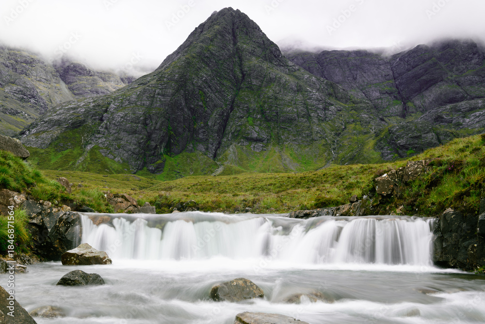 Fototapeta premium Waterfall and water stream, centered steep rocky mountain and ridge covered by clouds in background