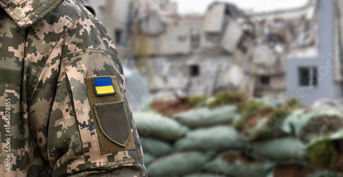 Photography Ukrainian soldier in military pixel uniform with a chevron of the Ukrainian flag against the background of a destroyed bombed building and defensive structures