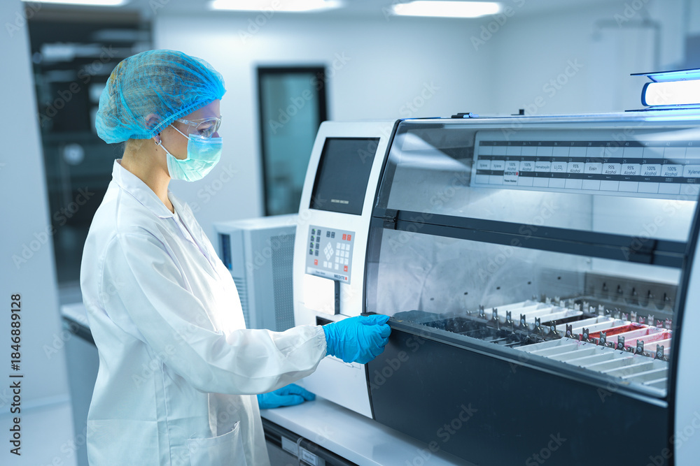 Naklejka premium Caucasian female scientist in full safety gear operating automated analyzer in sterile lab. Professional researcher setting machine parameters for medical biotechnology experiment.