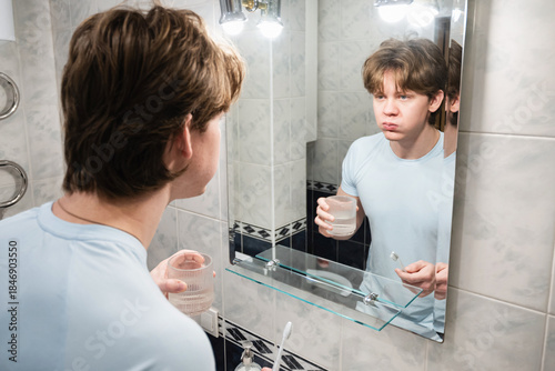 Slika na platnu Rinsing mouth with water from a glass, young adult caucasian man finishing oral hygiene routine in bathroom
