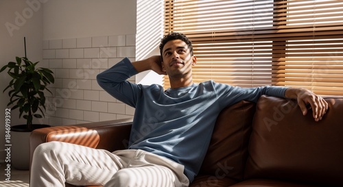 Young man relaxes on a brown leather sofa bathed in sunlight, enjoying leisure time