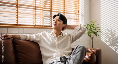 Young asian man relaxing comfortably on a leather sofa in sunlit modern room, enjoying rest