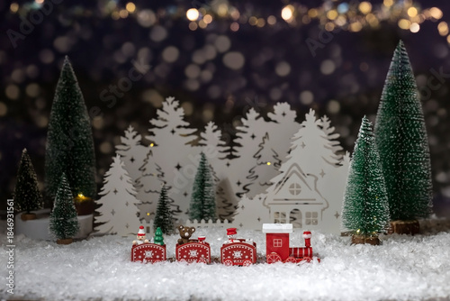 a toy train, a miniature Christmas house and fir trees on a table with artificial snow, blurred side lights in the background, a festive holiday scene.