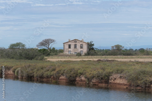 The photo shows an abandoned Mediterranean country house on the edge of a river, neglected and weathered, under a clear blue sky.