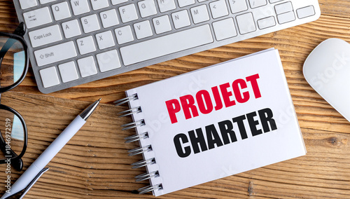 Notebook with text project charter on wooden desk with keyboard, pen, and glasses, symbolizing project planning