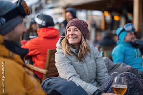 A cheerful woman in a winter jacket and knit hat smiles while holding a drink at a busy après-ski terrace with other skiers relaxing in the snowy alpine resort environment