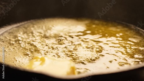 Close Up of a Thick Block of Yellow Butter Melting in a Black Cast Iron Skillet with Bubbling Golden Liquid and Tongs Gently Holding the Butter with Soft Dramatic Lighting