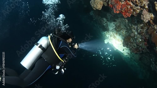 Solo Scuba Diver Explores Underwater Coral Reef Wall With Bright Flashlight Illuminating Dark Deep Blue Ocean Waters And Bubbles Rising Up From Equipment In Sunlit Water