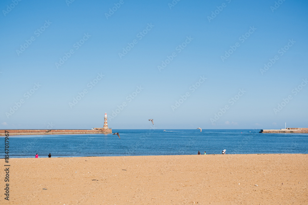 Fototapeta premium Sunderland UK: 16th July 2025: sandy Roker Beach in Sunderland. The sun shines brightly and boats can be seen on the water. A lighthouse stands in the distance.