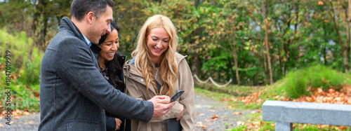 Portrait of diverse business colleagues stand outdoors in a park, smiling and laughing together while looking at smartphone during informal office break, enjoying a moment of connection and teamwork