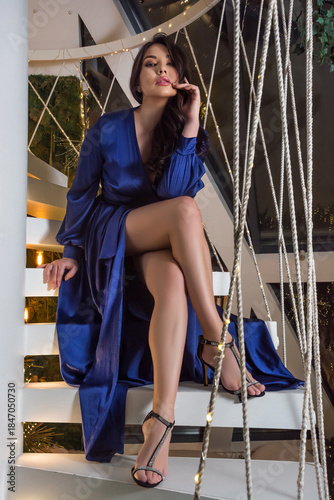 Beautiful woman is wearing blue satin dress and in high heels sits on the stairs at a christmas decorations