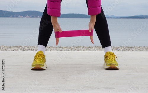 Outdoor resistance band exercise on beach in cold weather, front view, hands holding fitness band near legs, simple daily rehabilitation workout, recovery training, gentle fitness, wellness routine.