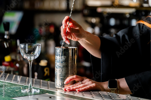 Close-up of a female bartender's hands mixing a cocktail in a silver shaker with a bar spoon at a dimly lit bar counter. The focus is on the drink preparation process