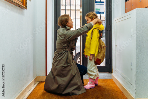 Valokuva Mother adjusting her daughter's yellow raincoat, preparing kid for school in hal