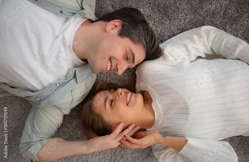 Young couple lying on a soft carpet at home, smiling and holding hands, enjoying a tender moment together. Cozy indoor lifestyle scene showing love, intimacy, trust, and emotional connection.