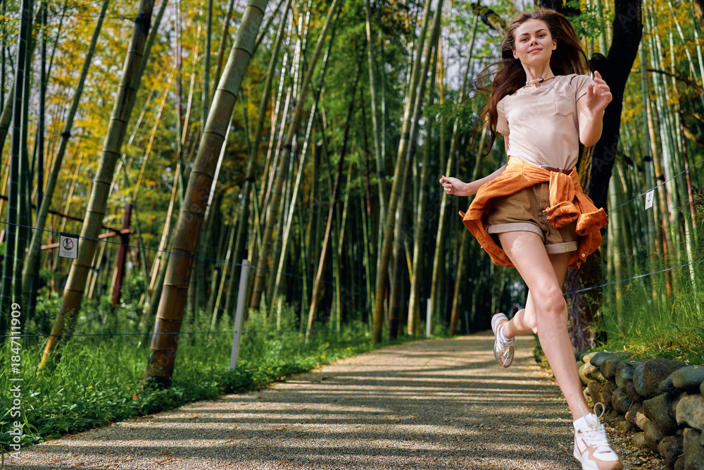 Fototapeta premium Woman jumping on a gravel path in a bamboo forest, casual outfit with sneakers, joyful happiness and motion captured during a summer walk on a green nature trail with sunlight.