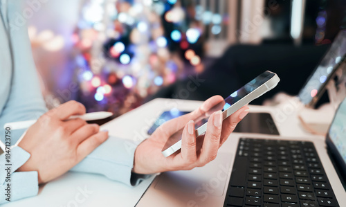 Woman using smartphone and digital pen on tablet near laptop during holiday season. Remote work or online shopping at night with blurred festive Christmas lights background