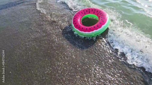 Vibrant swim ring in the shape of a refreshing slice of watermelon bobs in the ocean, being gently rocked by the waves under the warm sun