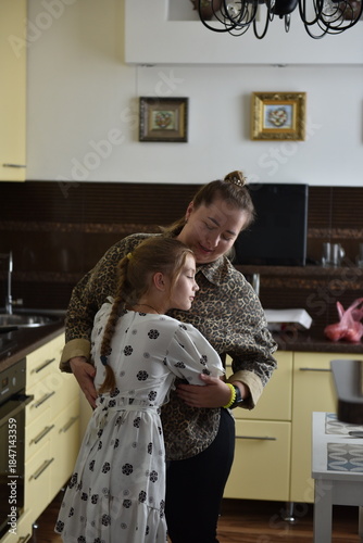 Adult woman hugging young daughter in home kitchen, tender family moment between mother and child, warm emotional connection, everyday family life, real people, authentic lifestyle scene