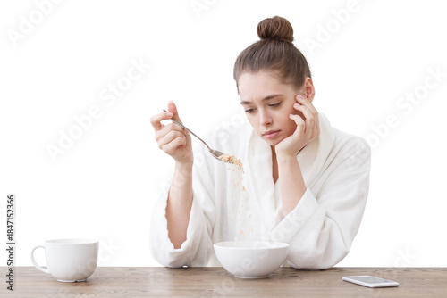 Young woman with no appetite, tired, bad mood and sleepy, sitting in the white loft kitchen, having unsavory unpalatable unappetizing breakfast