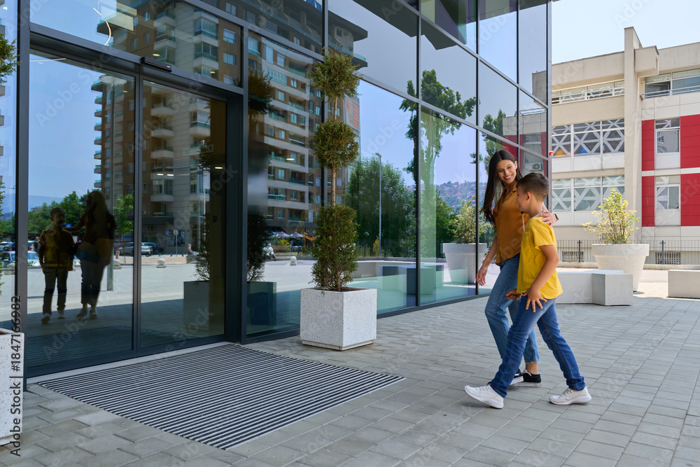 Fototapeta premium Mother and child walking towards a modern building entrance on a sunny day in a city environment