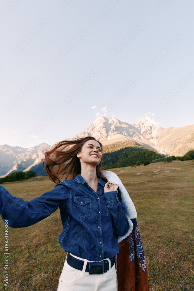 Naklejka premium Woman smiling in denim shirt on mountain meadow outdoors, enjoying nature and breeze. Portrait of happy traveler with jacket and open alpine landscape in soft sunlight.