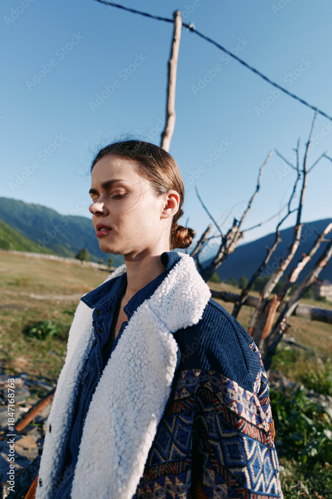 Naklejka premium Woman portrait outdoors in nature with mountains background, wearing patterned jacket and fleece coat, eyes closed in serene pose, sunlight on face and calm rural landscape mood