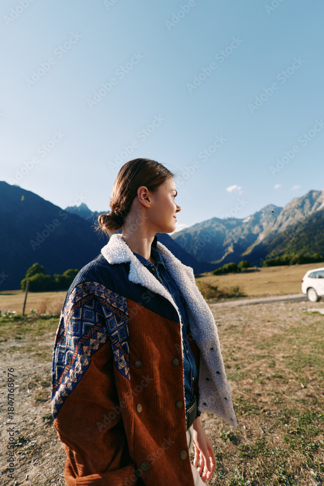 Naklejka premium Woman in profile wearing a patterned coat stands in countryside landscape with mountains in the background and a parked car, autumn travel scene with warm natural light and calm mood.