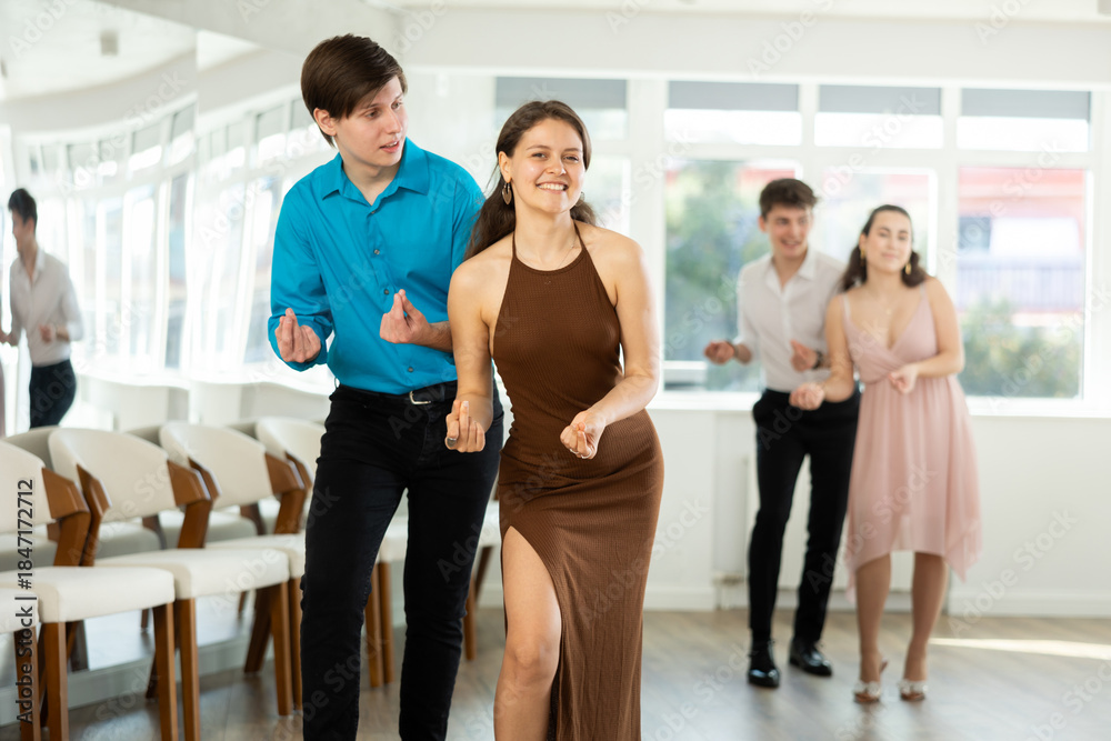 Fototapeta premium Smiling graceful girl in fitted brown dress and stylish guy in blue shirt and black trousers rehearsing jazz routine in dance studio, performing traveling steps and snapping fingers