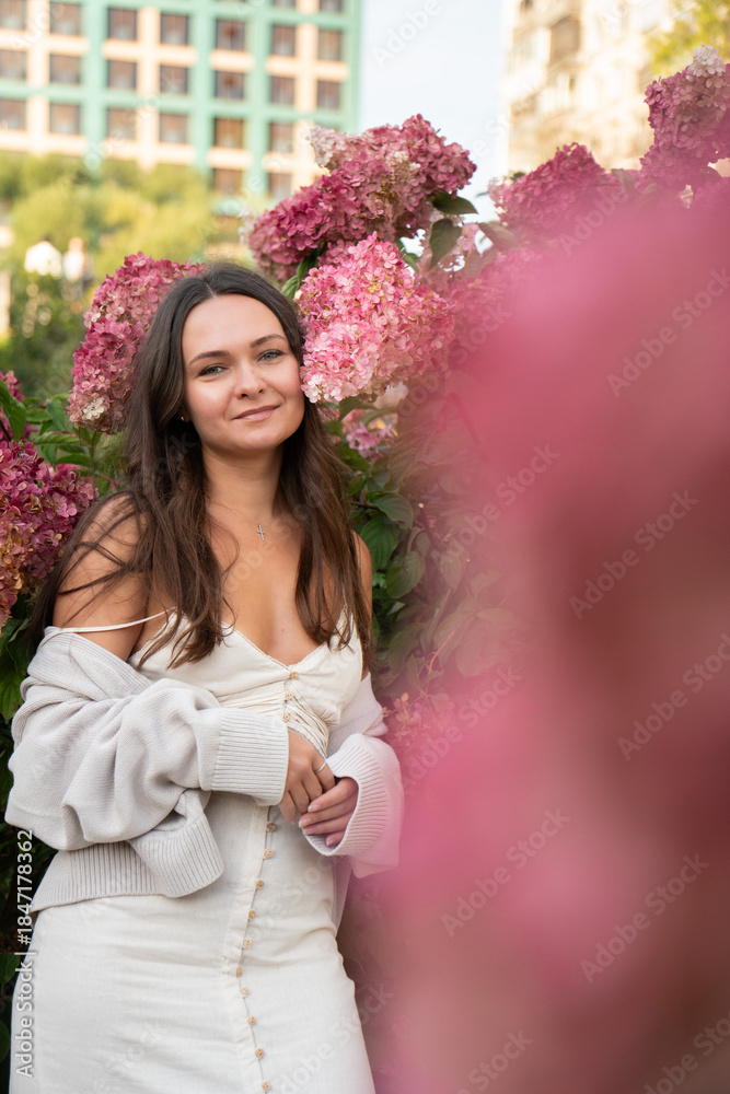 Fototapeta premium Young woman in a light dress stands among pink hydrangea flowers in an urban garden, soft daylight, pastel tones, calm and warm mood