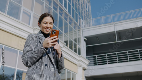 Woman in a gray suit smiles while using a smartphone outside a modern building on a sunny day