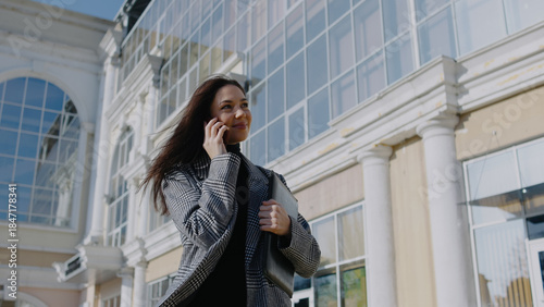 Woman speaks on phone while walking outside a building during daylight in an urban setting with large windows and columns