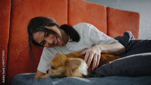 Woman smiles and pets small dog on bed in cozy room during morning hours while enjoying quiet time together