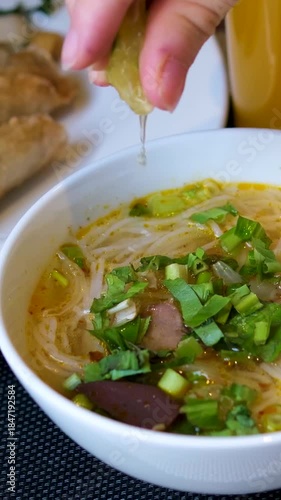 Woman hand squeezes fresh lime into traditional Vietnamese Pho noodle soup with beef, rice noodles, and herbs. Steam rises from the aromatic broth authentic taste of Vietnam.