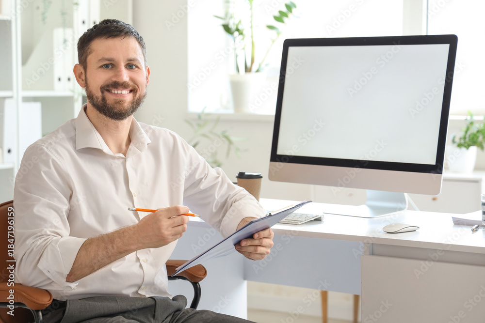 Naklejka premium Young businessman working with clipboard and blank computer at table in office