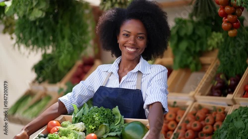 Young woman smiling at camera while holding a wooden crate overflowing with fresh organic vegetables and fruit, standing at a bustling farmers market stall, waist-up portrait