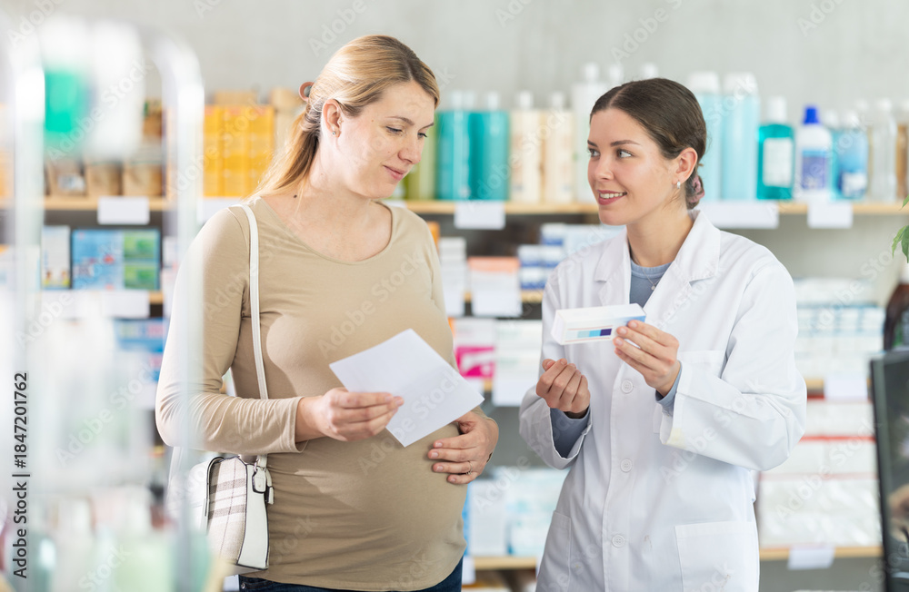Obraz premium Smiling young female pharmacist supporting expectant mother visiting pharmacy with prescription, recommending with care prenatal supplements best for pregnant women needs